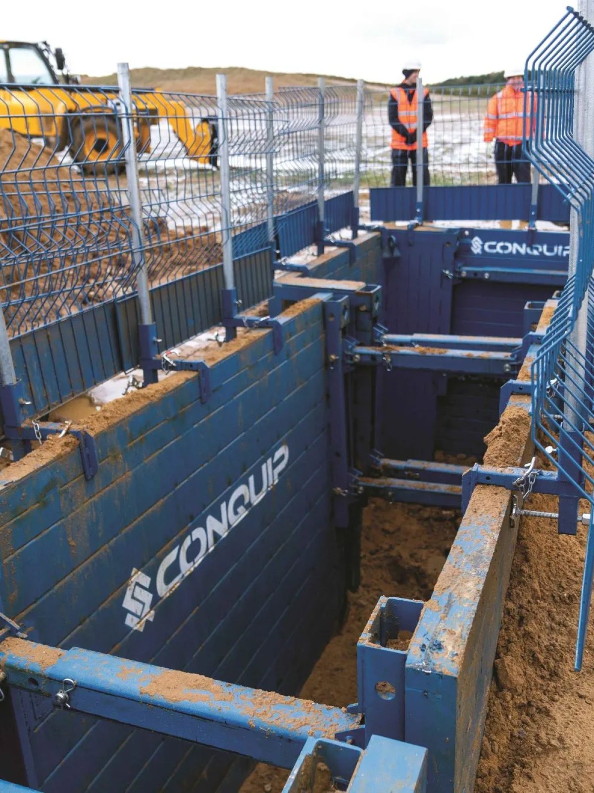 A trench supported by hydraulically strutted trench boxes, edge protection along both sides and operatives standing clear at the top